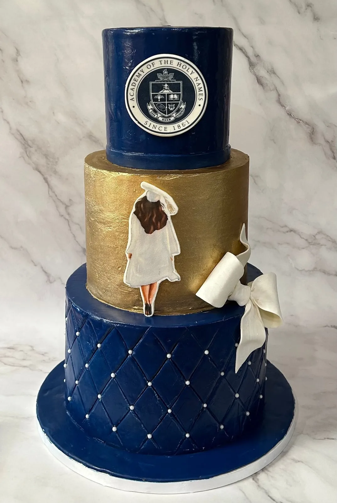 Three tier navy and gold graduation cake with a hand-painted Academy of the Holy Names crest and a sugar figure in cap and gown