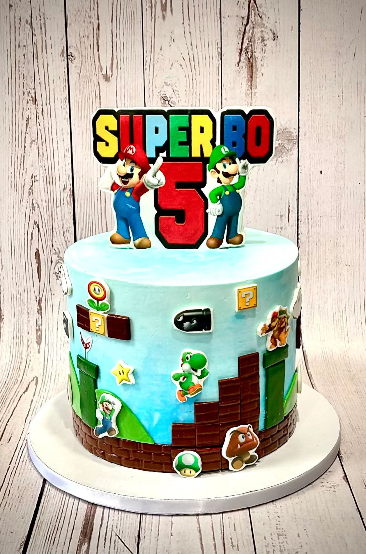 Super Mario fifth birthday cake with Mario and Luigi toppers, a painted buttercream sky and a fondant brick wall and pipes